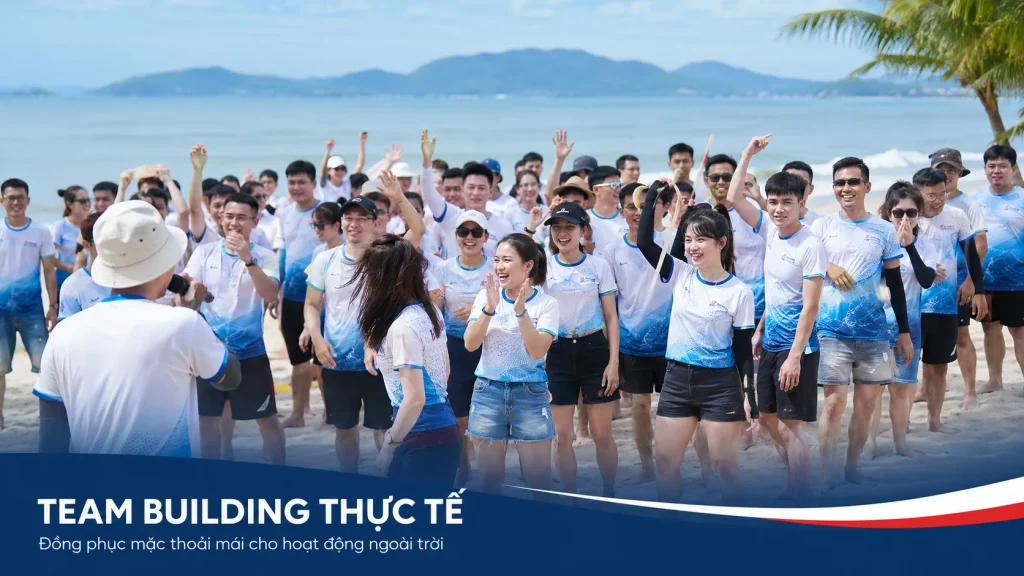 Ảnh team building thực tế trên bãi biển với đồng phục xanh trắng đồng bộ, thể hiện sự thoải mái khi hoạt động ngoài trời và tính nhận diện của áo team building.